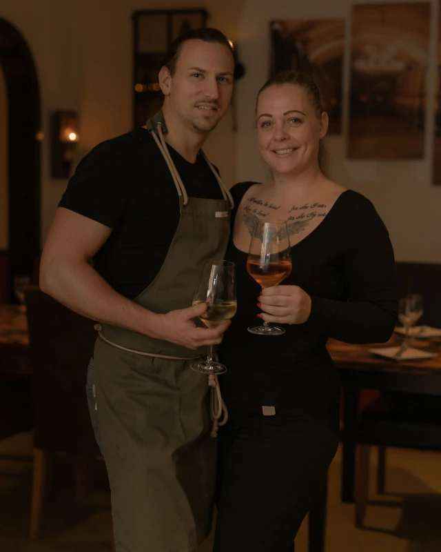 Florian_und_Kim Florian hat braune Haare, trägt eine grüne Schürze und hält in der rechten Hand ein Weißweinglas. Kim hat dunkelblonde, lange Haare, trägt schwarze Kleidung und hält in der linken Hand ein Roséweinglas.