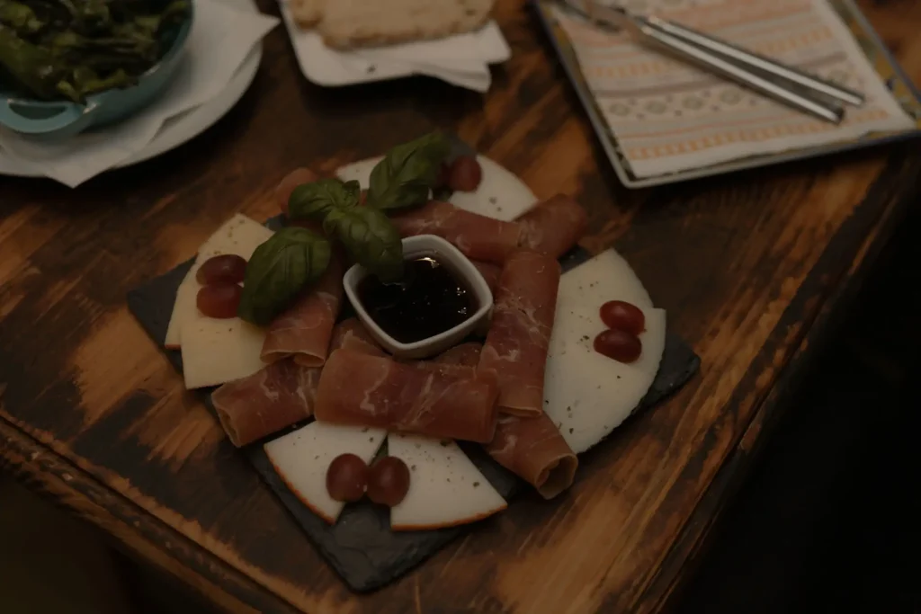 Auf einer Steinplatte wurden Käse-Spezialitäten mit Schinken, Weintrauben und Basilikum serviert. In der Mitte ist eine kleine Schale mit einem Dip.